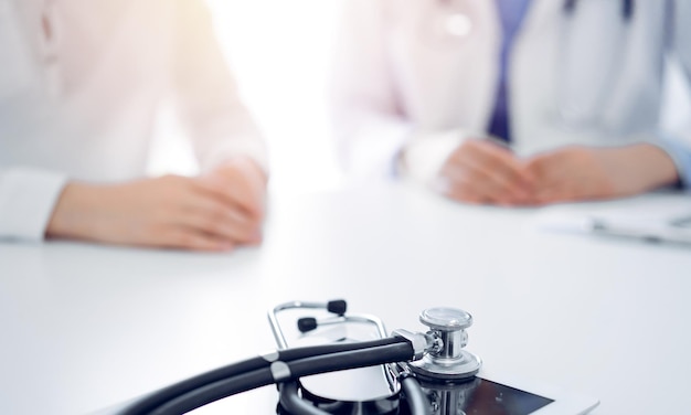 Stethoscope lying on the tablet computer in front of a doctor and patient sitting near of each other at the background. Medicine, healthcare concept.