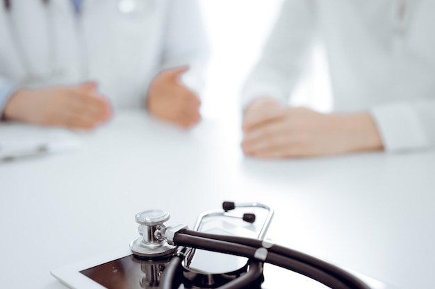 Stethoscope lying on the tablet computer in front of a doctor and patient sitting near of each other at the background. Medicine, healthcare concept.