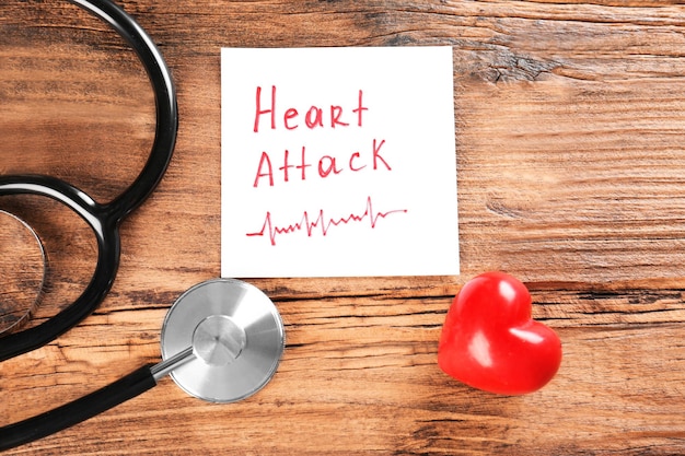 Примечание стетоскопа с фразой сердечный приступ и маленькое красное сердце на деревянном фоне