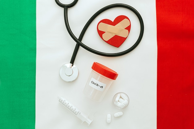 a stethoscope, pills, vaccine and a jar of coronavirus test over an Italian flag