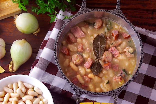 Stew or white bean soup with sausage, vegetables, spices and herbs served in rustic pan on wooden table