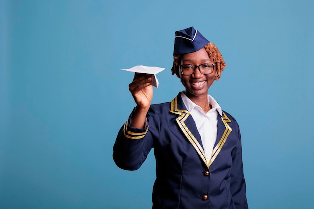 Stewardess in professionele uniform lancering papieren vliegtuigje voor camera in studio opname. Vrolijke vrouwelijke stewardess met bril en pet spelen tegen blauwe achtergrond.
