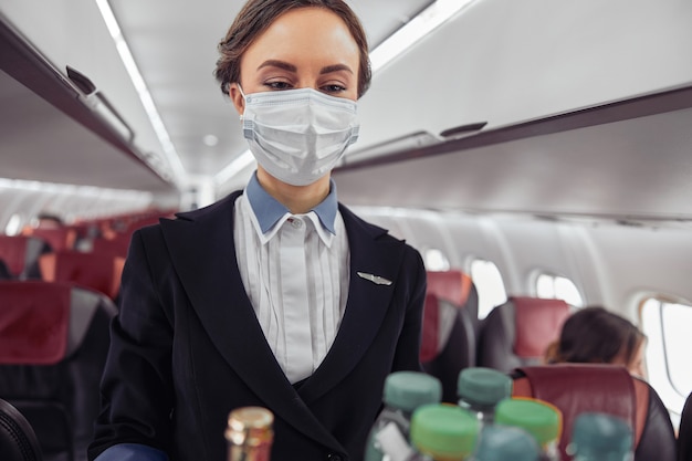 Stewardess met voedselkarretje in passagierscabine van vliegtuigjet. Moderne vliegtuig interieur. Europese vrouw draagt een uniform en medisch masker. Civiele commerciële luchtvaart. Vliegreisconcept