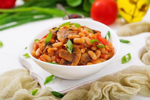 Stewed white beans with mushrooms and tomatoes with spicy sauce in a white bowl.