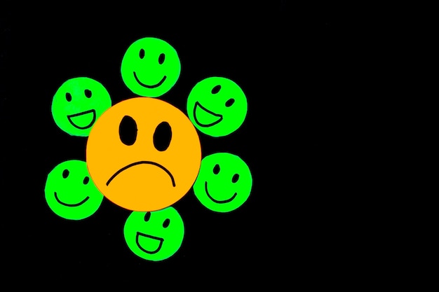 Photo stickers or paper cut of a sad yellow face surrounded by smiling and happy green faces on a black background