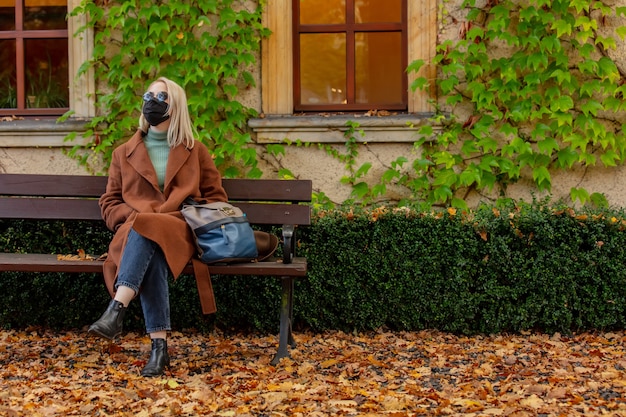 Stijl blonde in gezichtsmasker en jas zittend op een bankje in de herfst