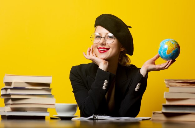 Stijl blonde vrouw in baret zittend aan tafel met boeken rond en houdt wereldbol op geel