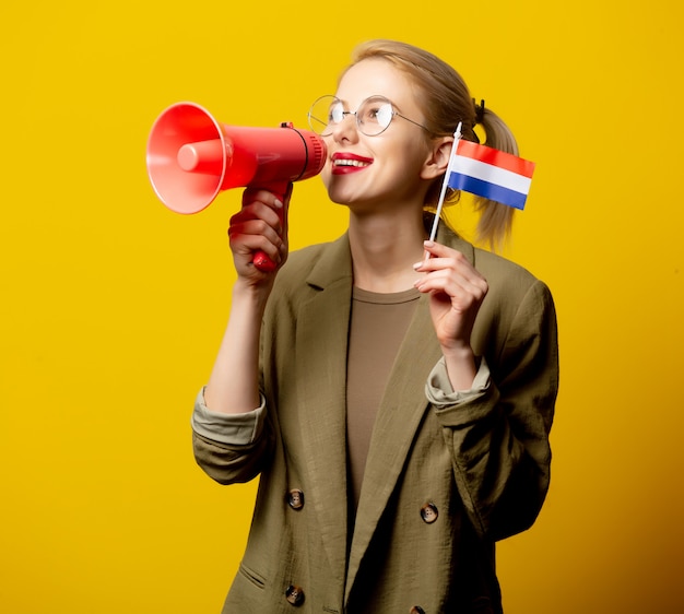 Stijl blonde vrouw in jas met Nederlandse vlag en megafoon op geel