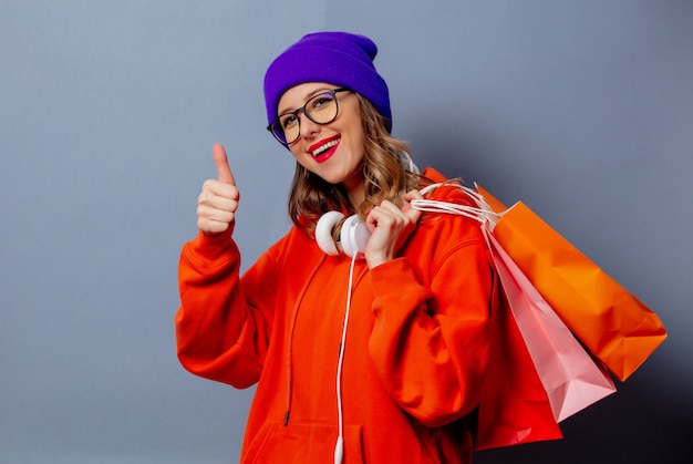 Stijlmeisje in oranje hoodie en purpere hoed met het winkelen zakken op grijze muur