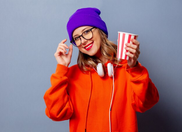 Stijlmeisje in oranje hoodie en purpere hoed met kop op grijze muur