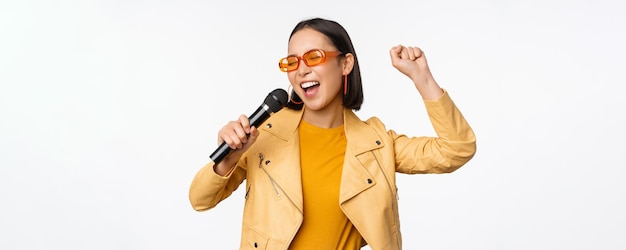 Stijlvol aziatisch meisje in zonnebril zingt liedjes met microfoon die microfoon vasthoudt en danst bij karaoke poseren tegen een witte achtergrond