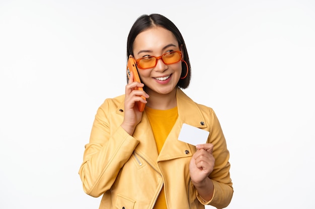 Stijlvol Aziatisch vrouwelijk model dat op een smartphone praat en een creditcard toont die over een witte achtergrond staat