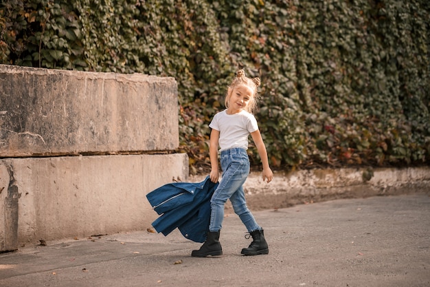 Stijlvol blond meisje in jeans en een wit T-shirt loopt door de straat. Meisje 7 jaar klein model, mooi kind