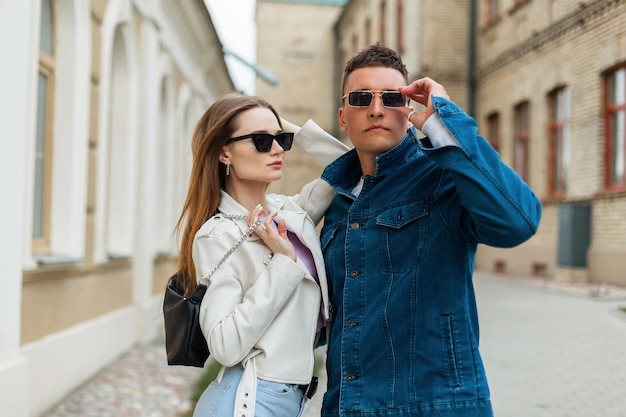 Stijlvol cool mooi paar modellen met trendy zonnebril in modieuze casual denimkleding en een modetas lopen op straat in de buurt van een vintage gebouw