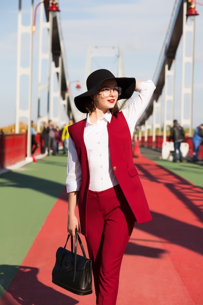Stijlvol geklede brunette vrouw in rood broekpak en zwarte hoed poseren op de brug