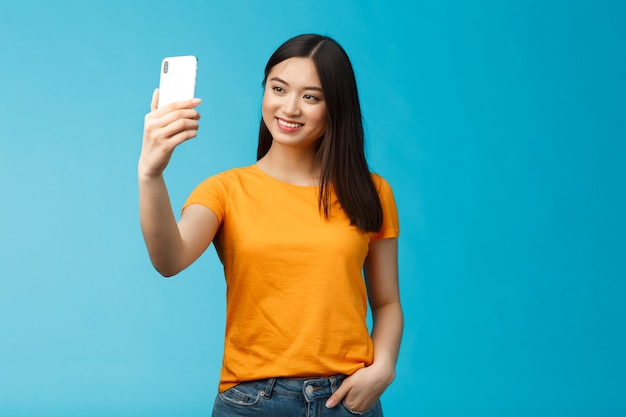 Stijlvol, goed uitziend aziatisch schattig meisje houdt smartphone vast en praat video-oproep glimlachend kijkt in grote lijnen naar het telefoonscherm, neemt selfie, voelt zich mooi, praat met volgers online sociale media, blauwe achtergrond.