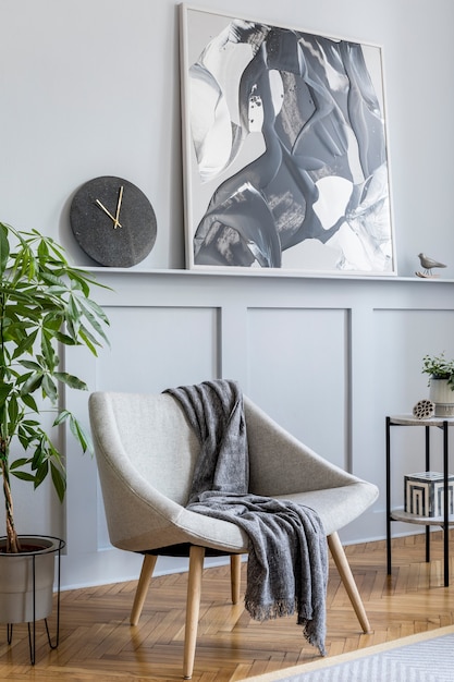 Stijlvol interieur van woonkamer met design grijze fauteuil, kussens, marmeren kruk, schilderijen, cactussen, decoratie, zwarte klok en elegante persoonlijke accessoires in modern interieur.