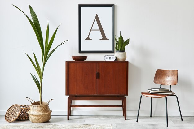 Stijlvol interieur van woonkamer met houten retro commode, stoel, tropische plant in rotan mand, decoratie en elegante persoonlijke accessoires. frame op de witte muur..