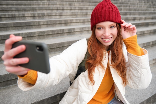 Stijlvol jong meisje met rode hoed neemt foto's op smartphonecamera en maakt selfie terwijl ze op trap n zit