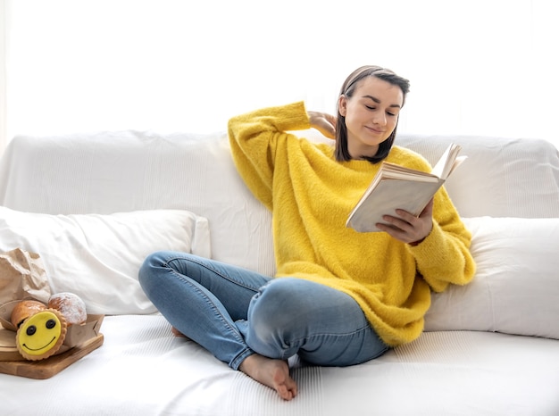 Stijlvol meisje in een gele trui rust thuis op de bank met een boek.