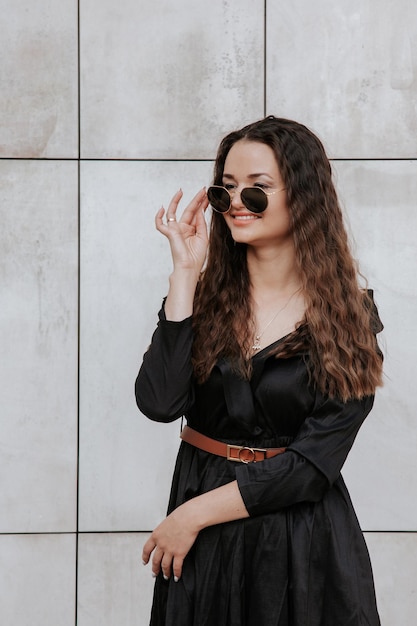 Stijlvol meisje in een zwarte jurk en zonnebril in de buurt van de grijze muur. Foto van vrouw in glazen. Jonge vrouw, krullend kapsel, glimlachend in de buurt van een muur in de straat. Meisje dat zwarte kledingskleding draagt.