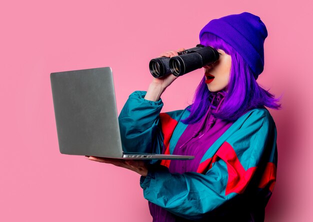 Stijlvol meisje in trainingspak kijken naar laptop met een verrekijker op roze muur