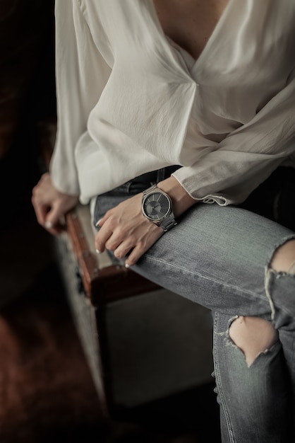 Stijlvol meisje met een horloge om haar pols gekleed in gescheurde spijkerbroek zit met gekruiste benen