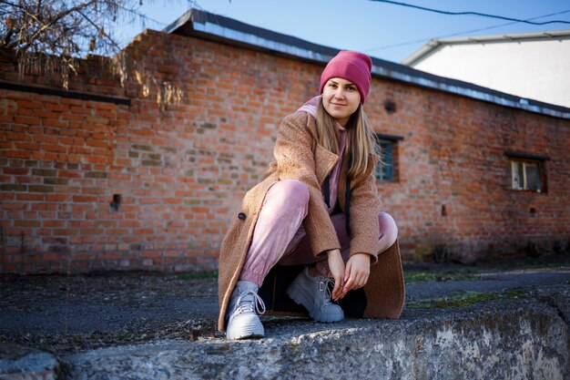 Stijlvol meisjesmodel in een bruine jas, roze pak en grijze laarzen op de stadsruïnes. De trends van de moderne mode. Modieus beeld