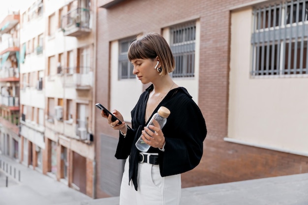 Stijlvol mooi europees meisje met kort kapsel met zwarte blouse en witte broek die smartphone gebruikt en muziek luistert terwijl ze buiten loopt Mooi meisje met een fles water