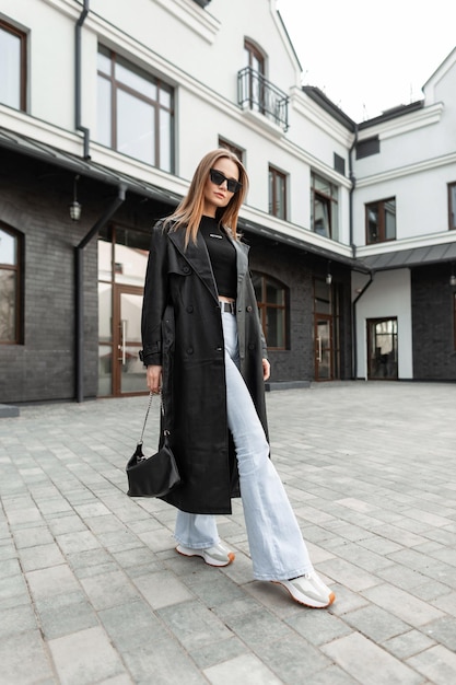 Stijlvol mooi jong meisje met zonnebril in modieuze outfit met een leren jas flare highwaisted jeans en sneakers met een portemonnee die op straat loopt