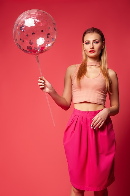 Stijlvol, mooi jong vrouwelijk model met lichte make-up met een roze top en rok die een transparante ballon met confetti vasthoudt terwijl ze tegen een rode achtergrond staat