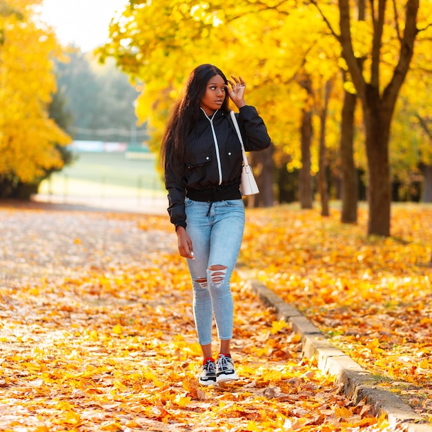 Stijlvol mooi jong vrouwenmodel in vrijetijdskleding loopt in een herfstpark met felgekleurd bladgoud