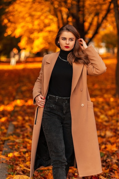 Stijlvol mooi zakenmeisje met rode lippen in een modieuze beige jas met een trui en zwarte spijkerbroek loopt in een park met gekleurd herfstgebladerte