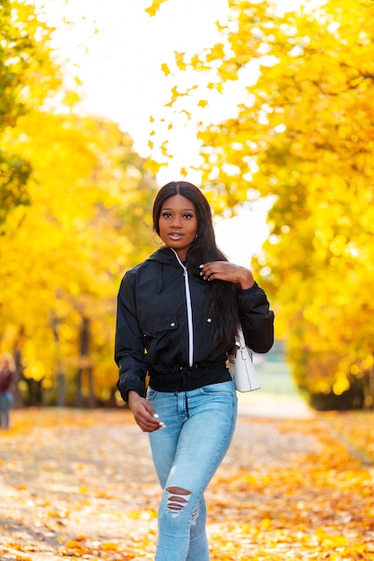 Stijlvol mooi zwart meisje in modieuze kleding met een casual jas en spijkerbroek met een handtas loopt in een herfstpark met kleurrijke gouden herfstbladeren