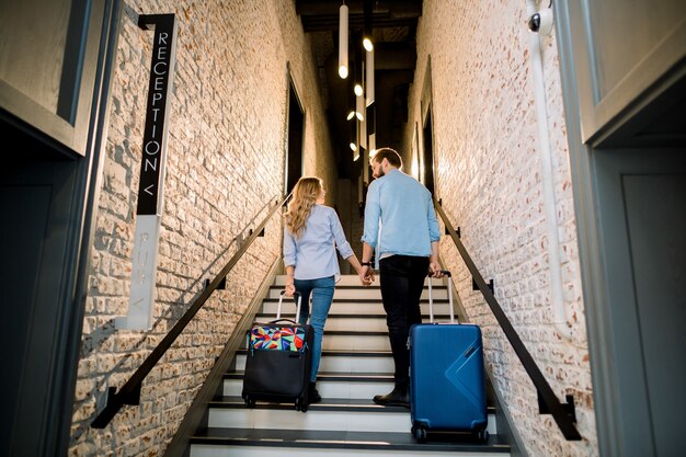 Stijlvol paar toeristen hand in hand en koffers trekken, man en vrouw in vrijetijdskleding, lopen naar boven, terwijl ze aankomen in het hotel. Zakenreis, vakantie