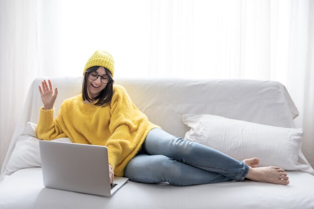 Stijlvol vrolijk brunette meisje in glazen en een hoed ligt op de Bank voor een laptop en communiceert online kopie ruimte.