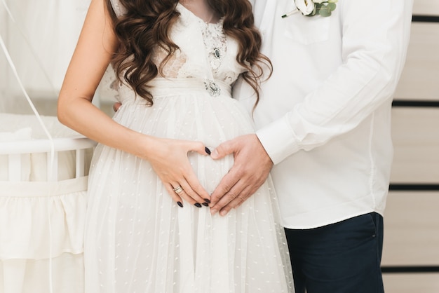 Stijlvol zwanger meisje in witte kleren en naast haar man omhelst haar buik en houdt haar handen in de vorm van een hart