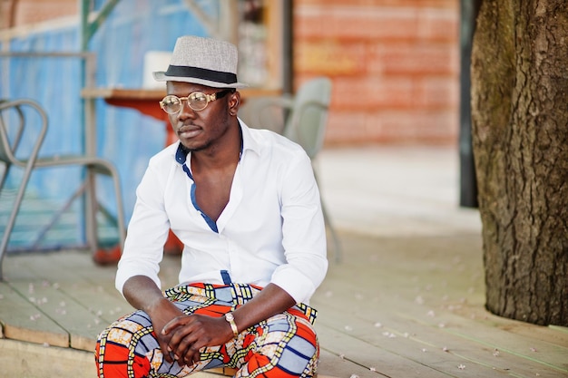 Stijlvolle Afro-Amerikaanse man in wit overhemd en gekleurde broek met hoed en bril poseerde buiten Zwarte modieuze modeljongen