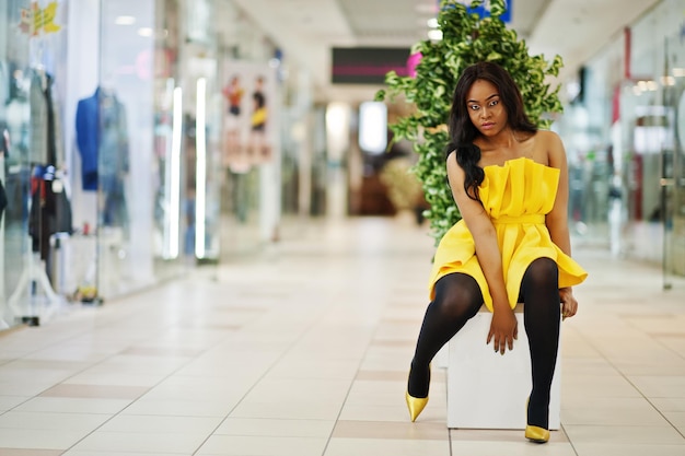 Stijlvolle Afro-Amerikaanse vrouw in gele jurk poseerde in het winkelcentrum en zat binnen op de bank