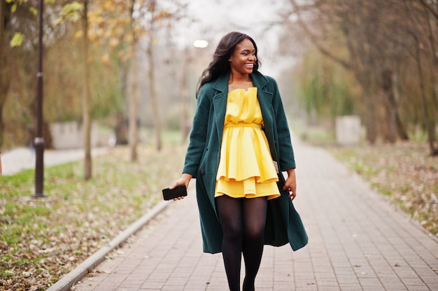 Stijlvolle afro-amerikaanse vrouw op groene jas en gele jurk gesteld tegen herfst park met mobiele telefoon op handen.