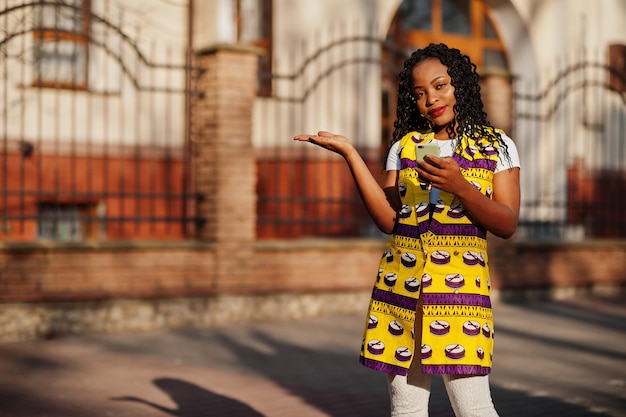 Stijlvolle Afro-Amerikaanse vrouwen in gele jas poseren op straat op zonnige dag met mobiele telefoon bij de hand