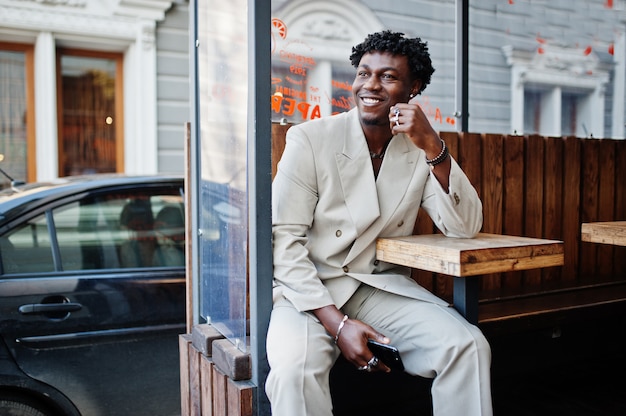 Stijlvolle afro man in beige oude school pak zitten aan tafel op straat. Modieuze jonge Afrikaanse man in casual jas op blote romp.