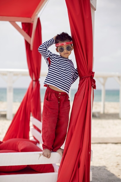 Stijlvolle babyjongen in een gestreept T-shirt en zonnebril staat op de rode strandzonnebank