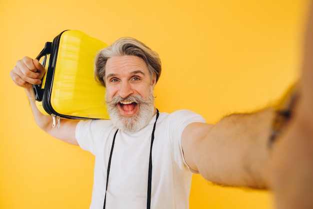 Stijlvolle bebaarde en besnorde man glimlachend en het nemen van een selfie op zijn smartphone met een gele koffer.