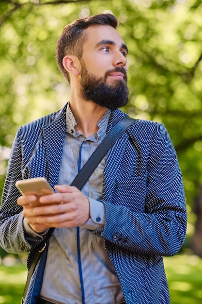 Stijlvolle bebaarde man in een blauwe jas met smartphone in een park.