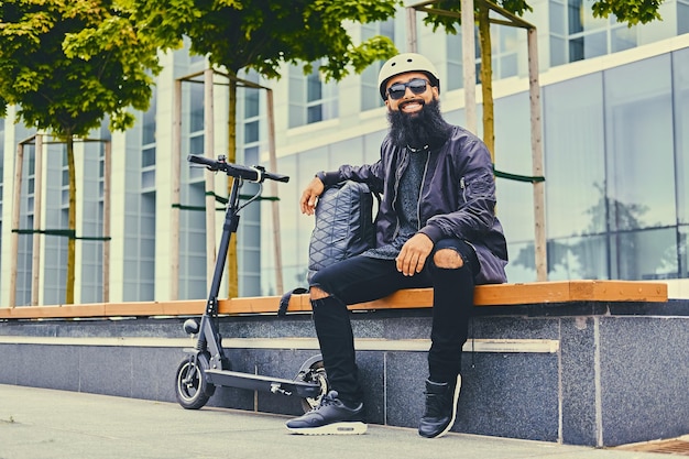 Stijlvolle bebaarde man in zonnebril ontspannen op een bankje na het rijden met een elektrische scooter in het centrum.