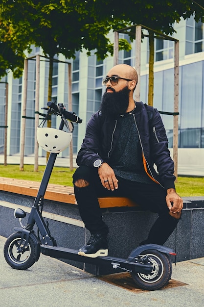 Stijlvolle bebaarde man in zonnebril ontspannen op een bankje na het rijden met een elektrische scooter in het centrum.