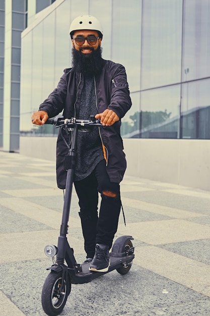 Stijlvolle bebaarde man in zonnebril poseren op elektrische scooter in meer dan modern gebouw achtergrond.