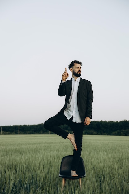 Stijlvolle bebaarde man in zwarte jas en wit overhemd poseren voor een foto in het midden van een groen tarweveld