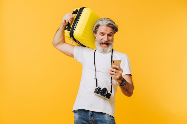 Stijlvolle bebaarde man lachen, met een gele koffer en kijken naar de telefoon die een land kiest om te reizen.